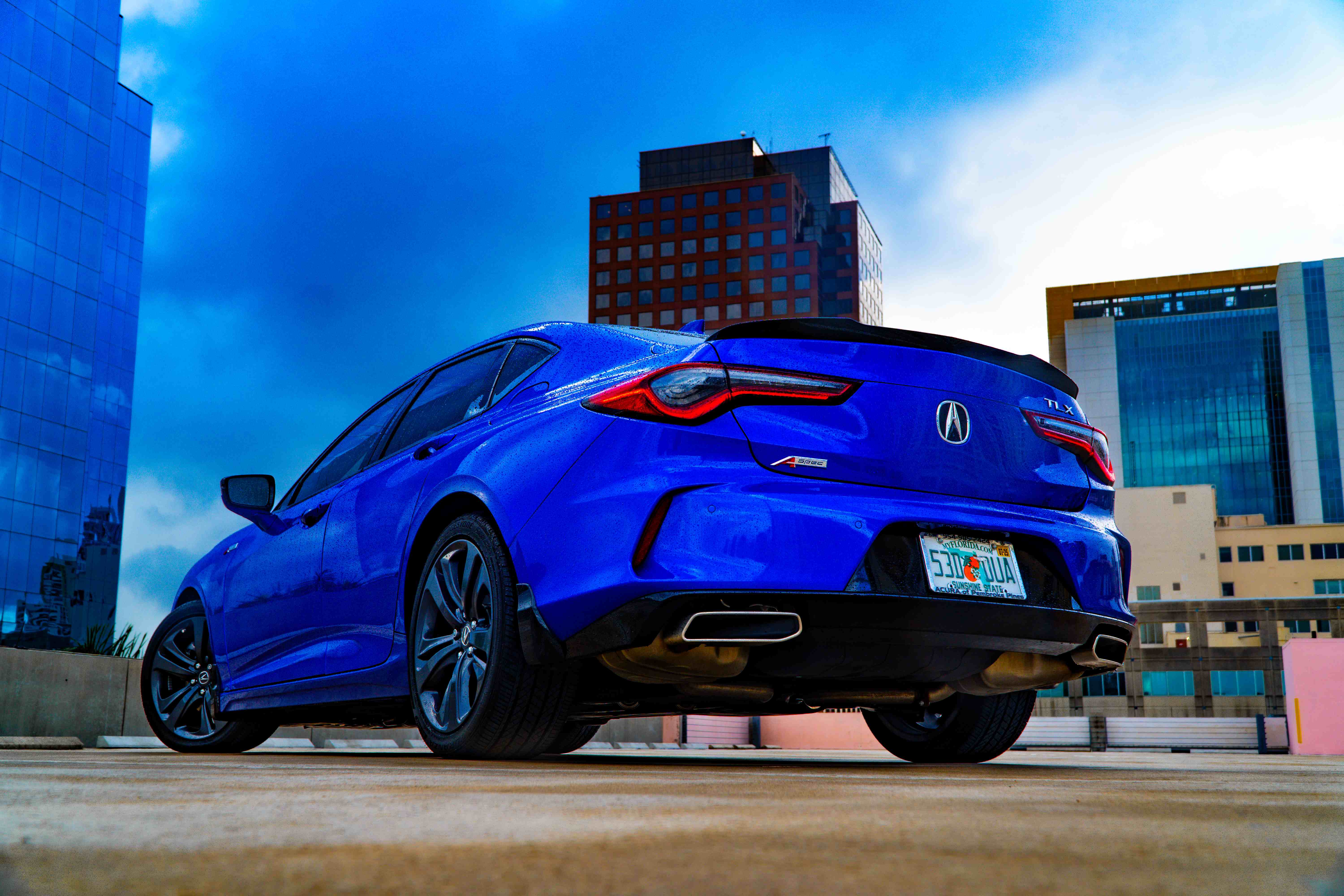 Low-angle rear three-quarter view of a blue Acura TLX A-Spec on a rooftop parking deck, framed by a glass tower and red brick high-rise under a deep blue sky