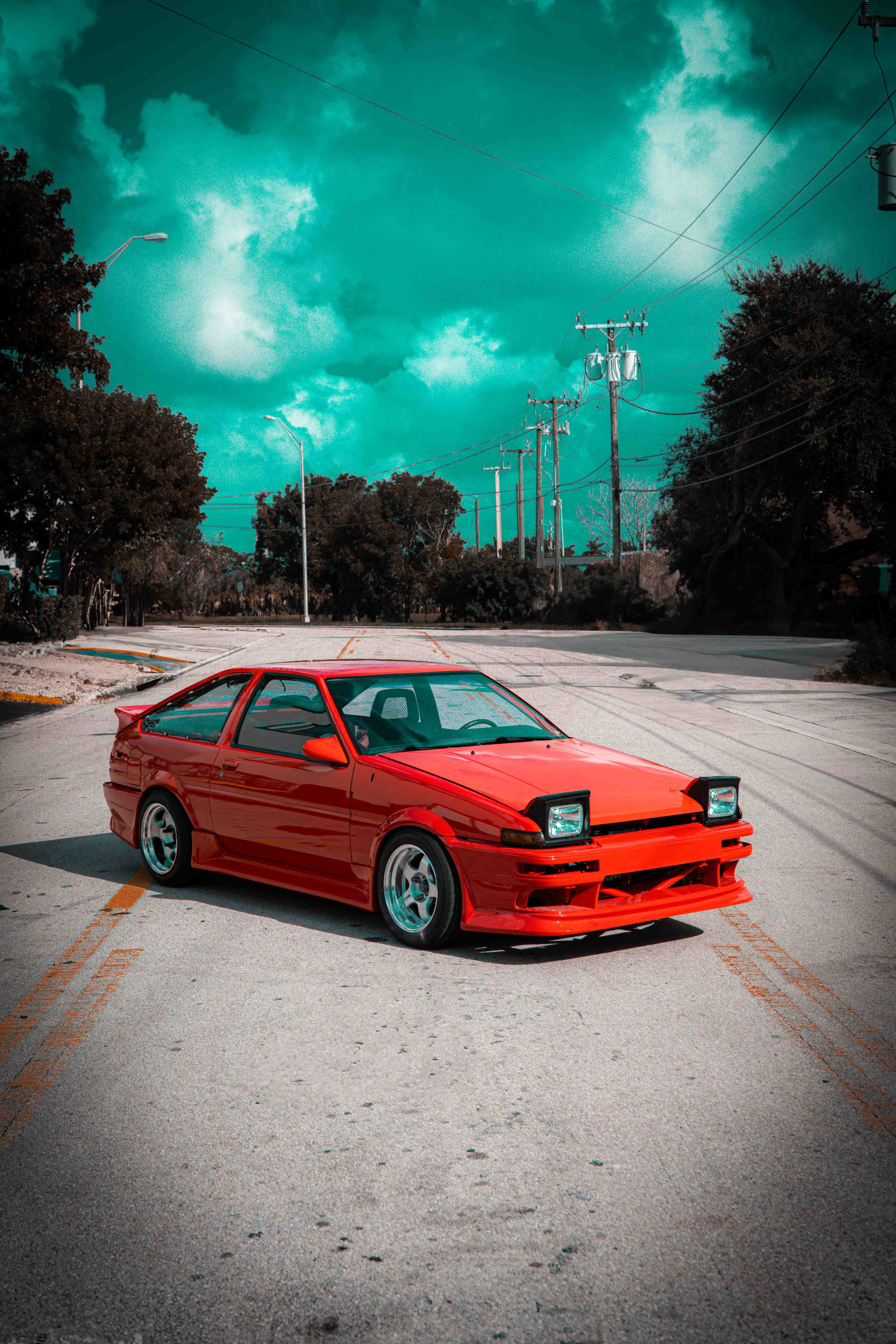 Red Toyota AE86 Corolla hatchback parked in the middle of an empty street under dramatic teal clouds, lined by trees and power poles