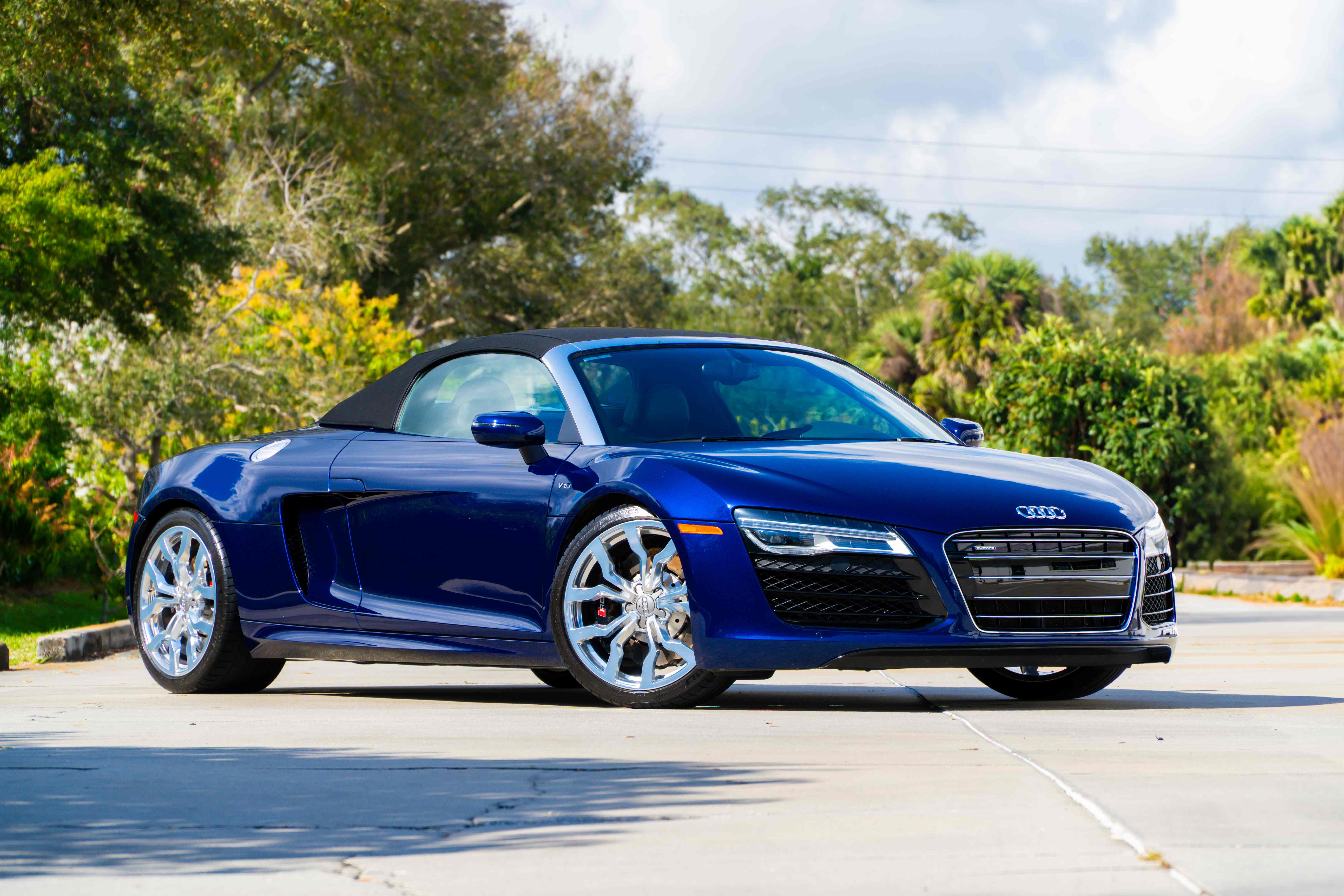 Front three-quarter view of a blue Audi R8 V10 Spyder with its black soft top up and polished wheels, parked on concrete in front of lush trees