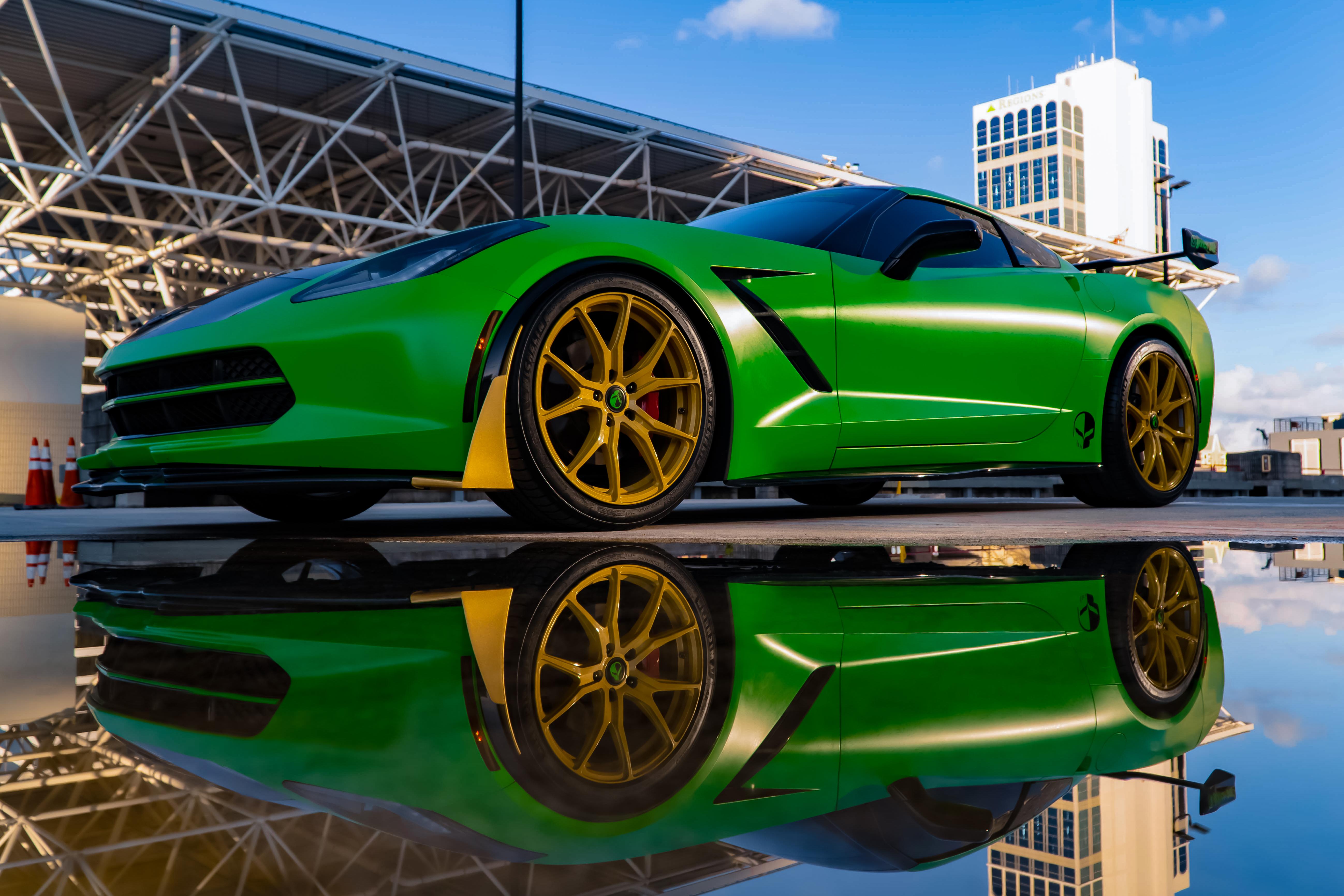 Low front three-quarter view of a green Corvette C7 with gold wheels reflected in a rooftop puddle beneath a steel truss canopy