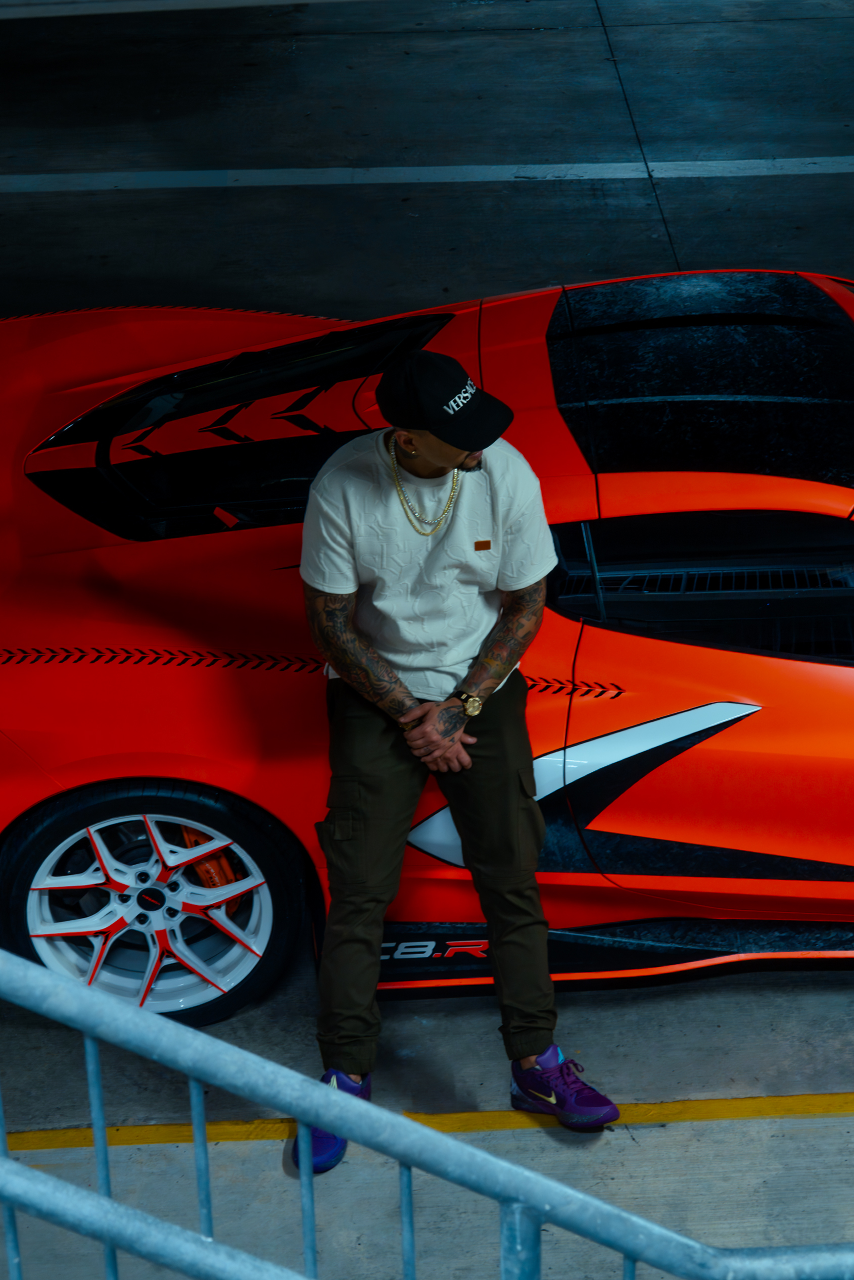 Overhead view of a man in a white tee and Versace cap leaning against the side of the orange Corvette C8 with white and red wheels