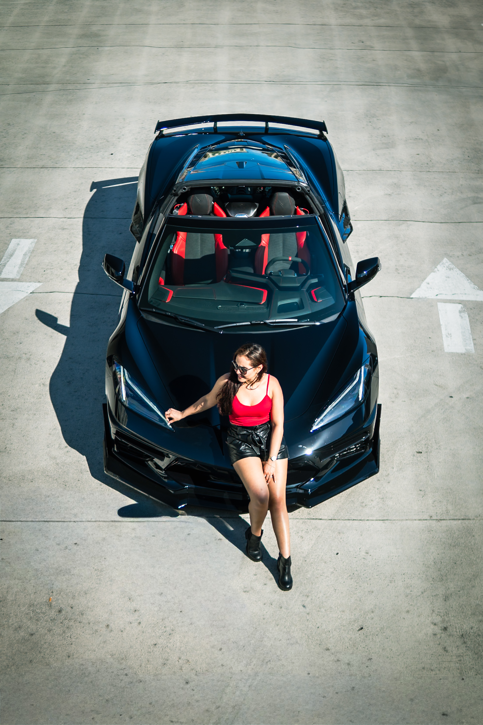 Overhead view of a woman in a red top sitting on the hood of the black Corvette C8 convertible with red seats