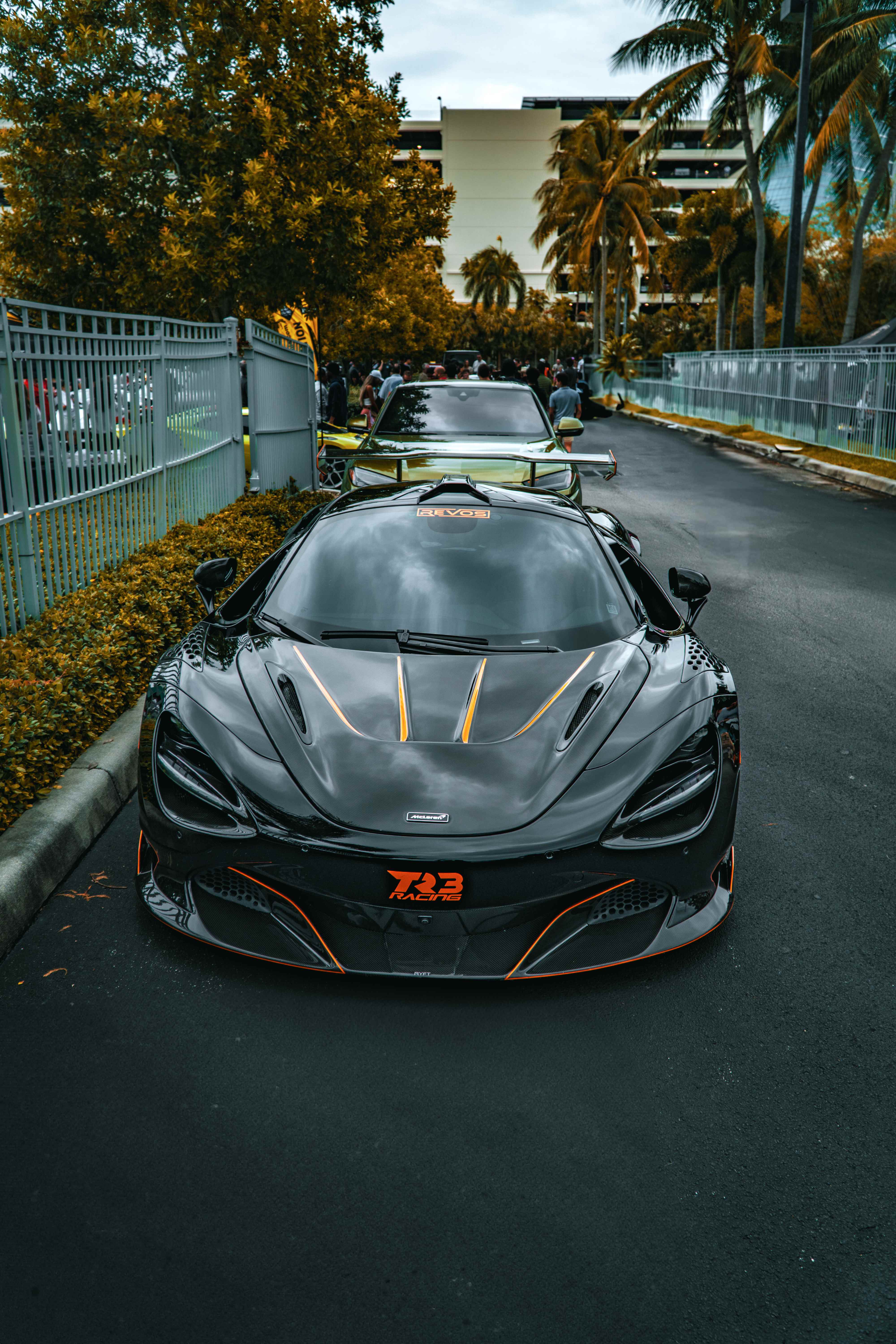 Head-on view of the black McLaren with orange accents and TR3 Racing badge at a car meet lined with palm trees