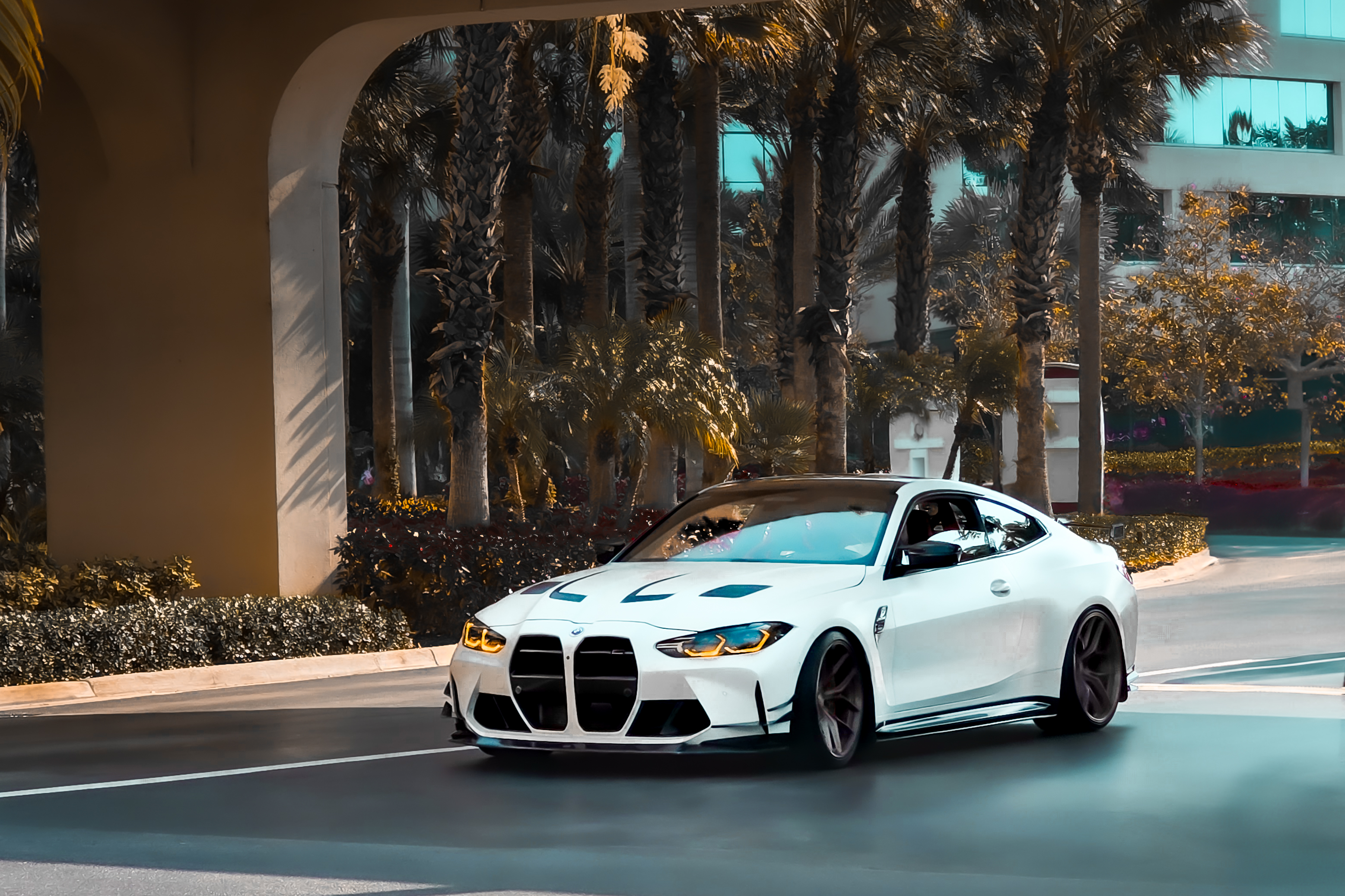 White BMW M4 with amber headlights driving past an arched overpass lined with palm trees