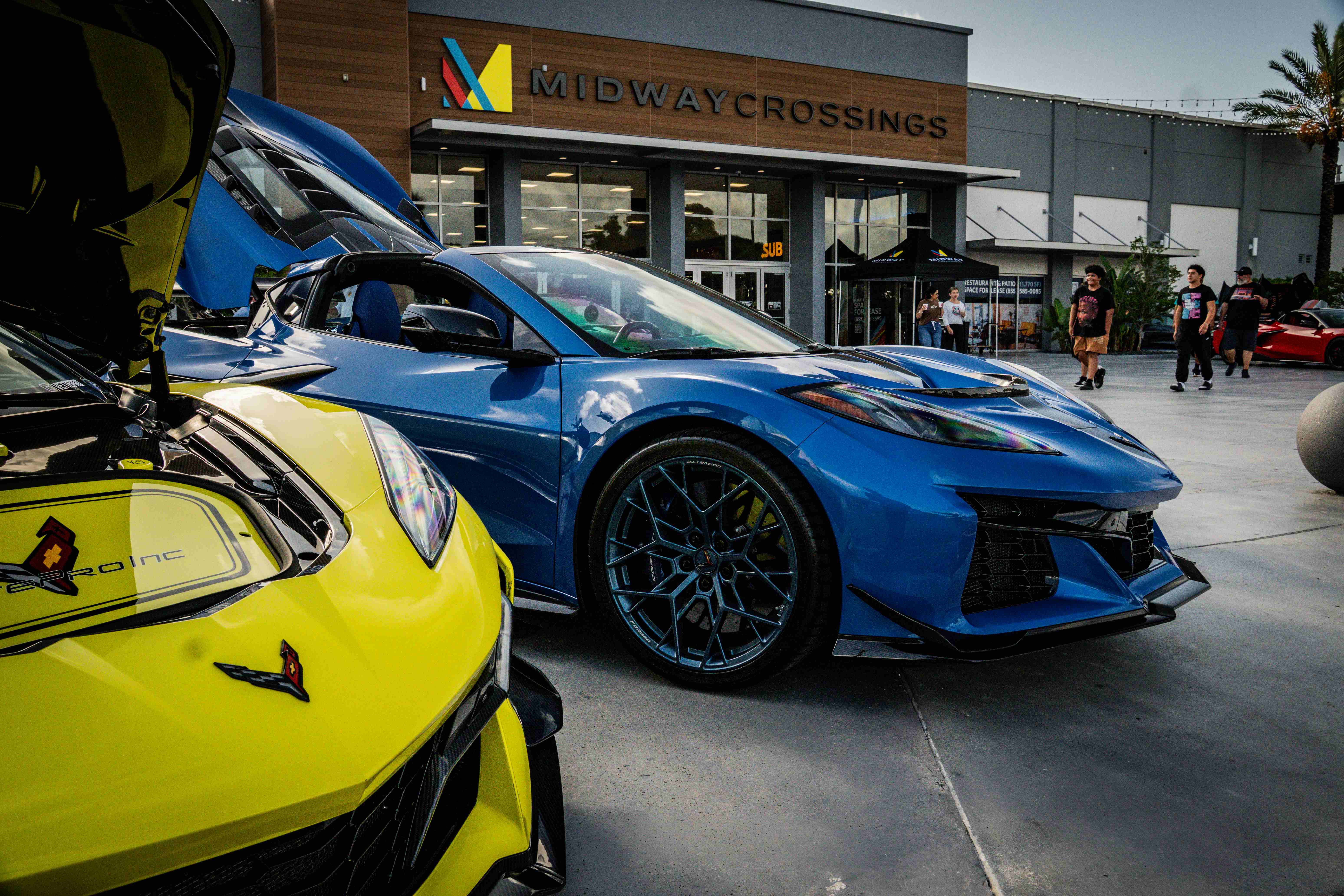 Blue Corvette C8 Z06 with doors raised beside a yellow VettePro Corvette in front of Midway Crossings